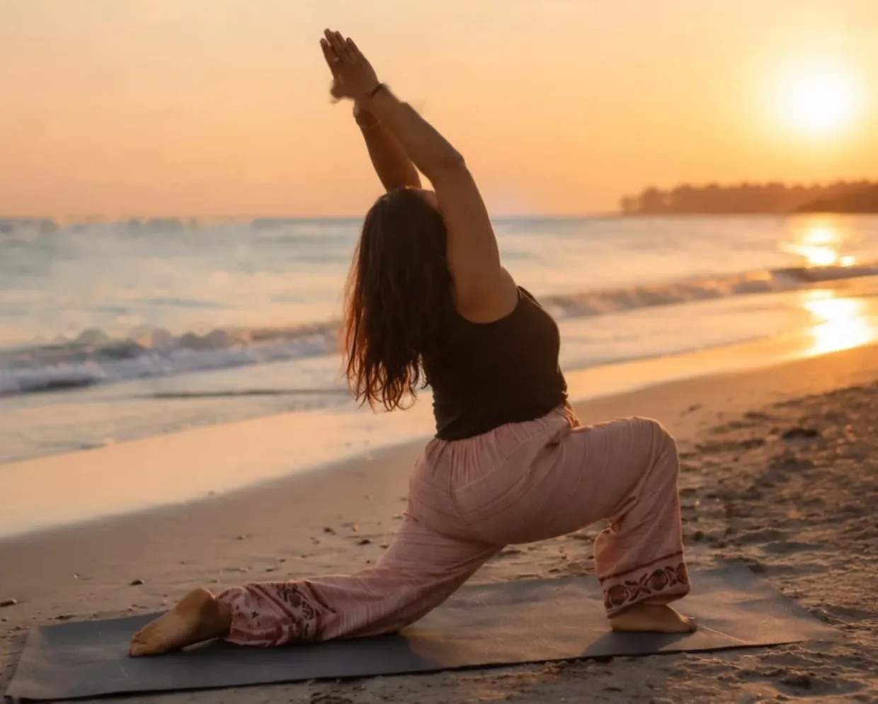 Woman in a low lunge with arms raised, practising yoga on a beach at sunset