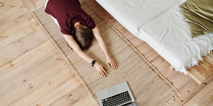 Overhead view of a person in child pose on a jute rug beside a bed, with a laptop on the wooden floor next to them