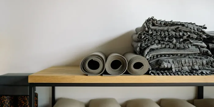 Rolled grey yoga mats and a stack of folded striped blankets on a wooden shelf, with bolsters underneath