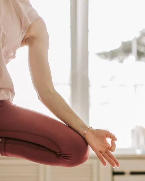 A person in rose-coloured leggings seated in a mudra by a bright window, face out of frame