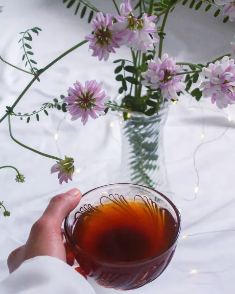 A hand holding a glass cup of dark tea beside a vase of pale purple wildflowers on white linen