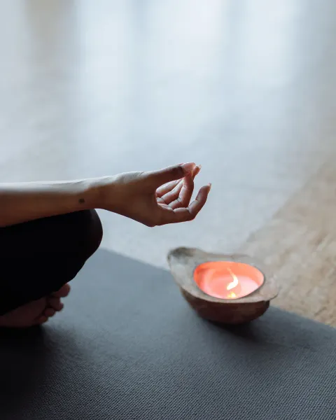 A hand resting in a mudra on a knee beside a small lit candle in a clay bowl, on a dark mat